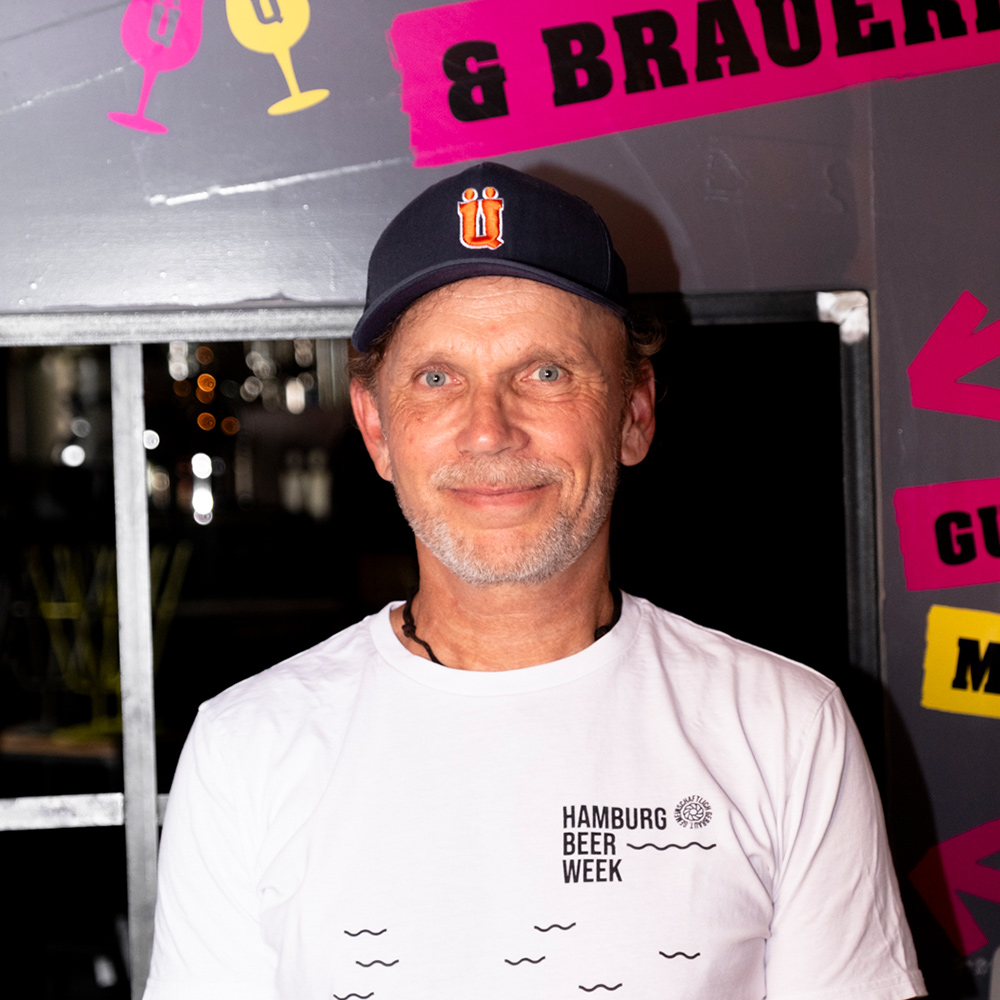 Axel Ohm von ÜberQuell trägt T-Shirt und Kappe der Hamburg Beer Week, bunter Festivalhintergrund.