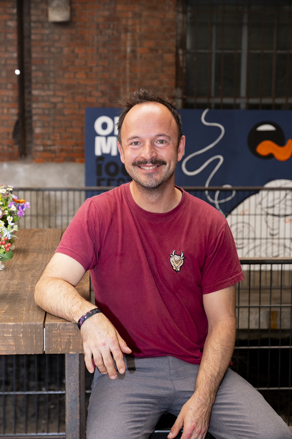 Thomas Sampl von der Hobenköök sitzt lächelnd am Holztisch vor OPEN MOUTH Festivalbanner.
